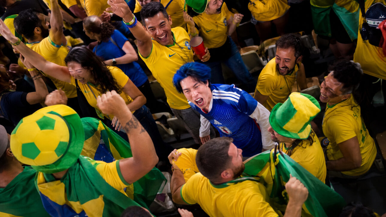 Torcedor japonês de cabelo azul grita entre torcedores brasileiros com camisas amarelas e bandeiras do Brasil em um estádio.