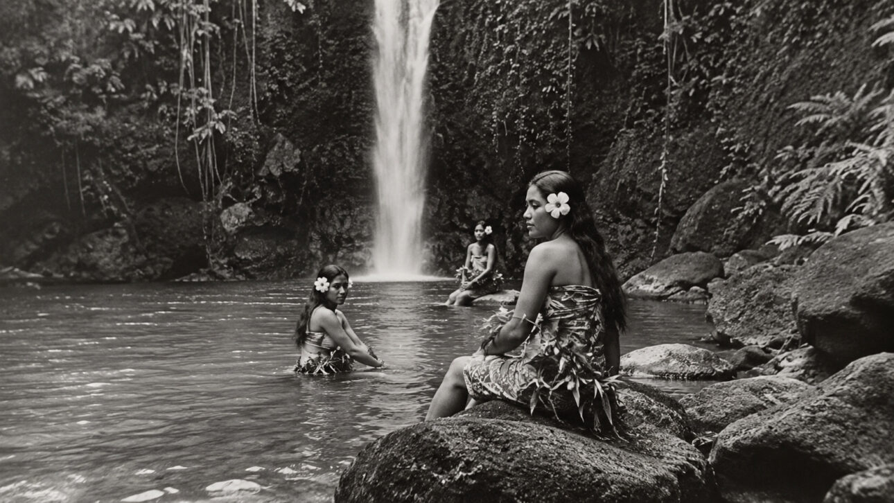 Samoan women in a lagoon surrounded by stones and vegetation, in front of a waterfall, in an image inspired by the 1926 documentary Moana.