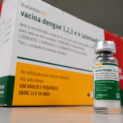 Frasco da vacina Butantan-DV contra a dengue em destaque diante da embalagem do imunizante, representando a suspensão temporária da vacinação após investigação de eventos adversos no Brasil.
