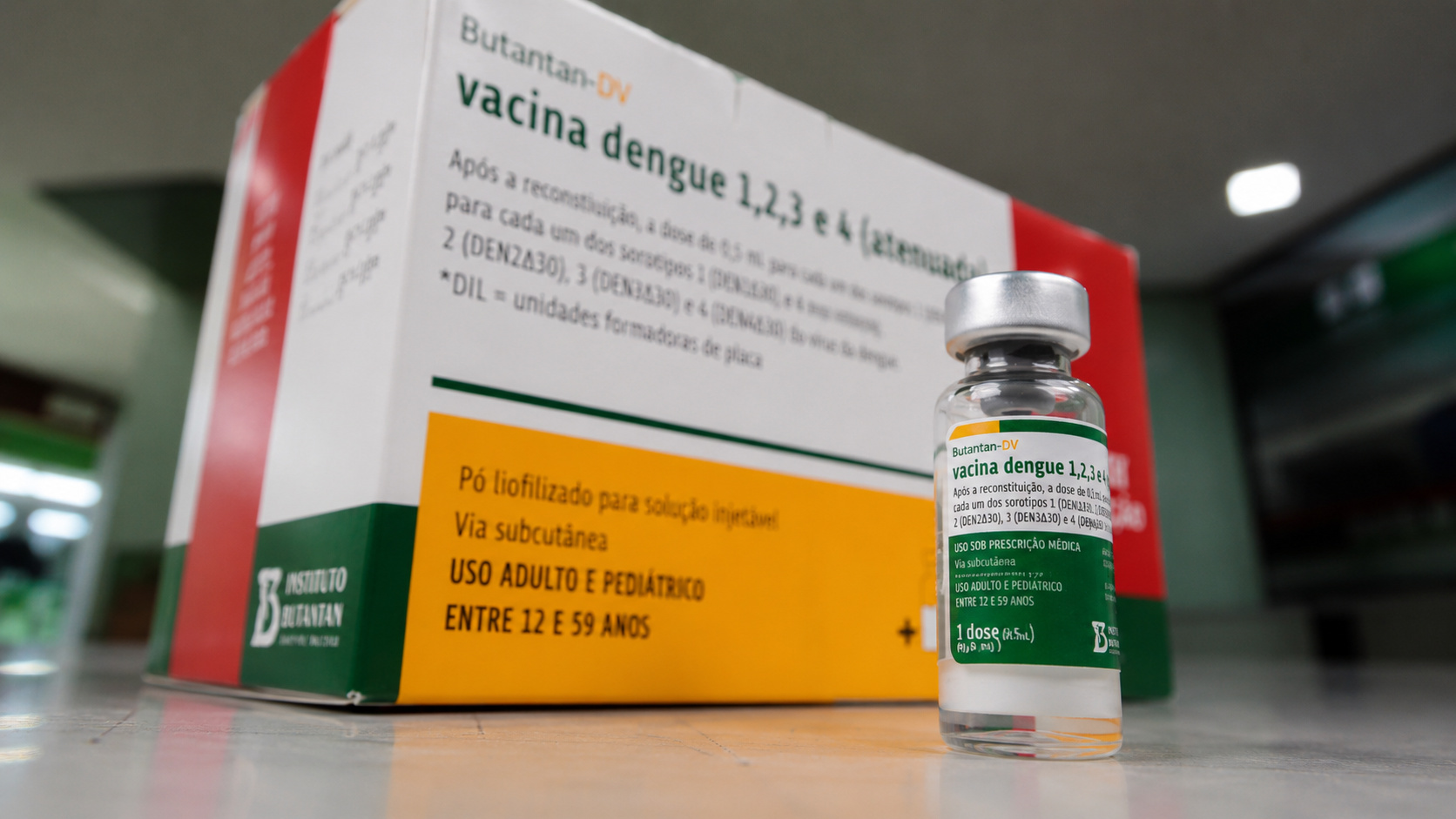 Frasco da vacina Butantan-DV contra a dengue em destaque diante da embalagem do imunizante, representando a suspensão temporária da vacinação após investigação de eventos adversos no Brasil.