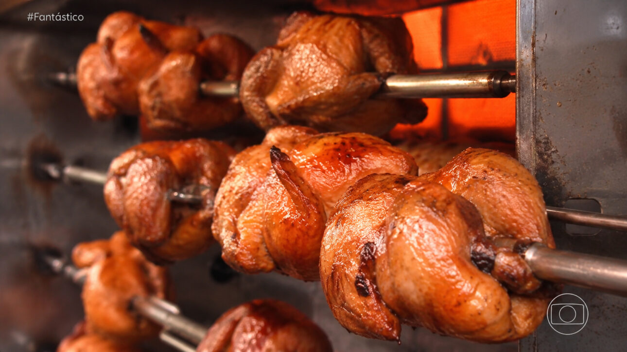 Frangos assados giram em equipamento de rotisserie industrial, representando produtos alimentícios comercializados por comerciantes afetados pelo controle de fornecedores em comunidades do Rio de Janeiro.