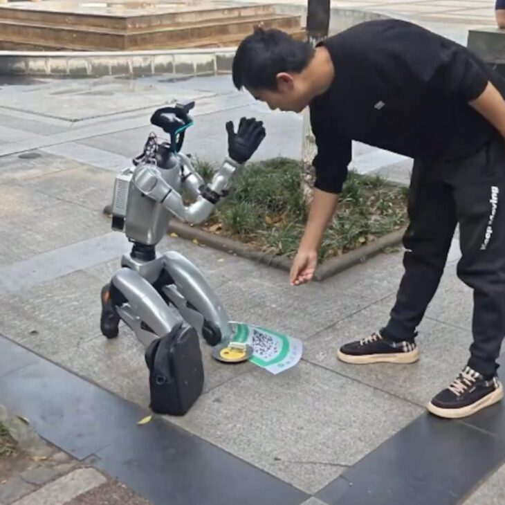 Robô humanoide de aparência semelhante a modelos comerciais chineses aparece ajoelhado em uma calçada com QR Code e tigela para doações; equipamentos desse tipo, como o Unitree G1, podem custar a partir de US$ 13,5 mil, o que reforça a dúvida sobre se a cena seria marketing, sátira social ou performance sobre inteligência artificial.