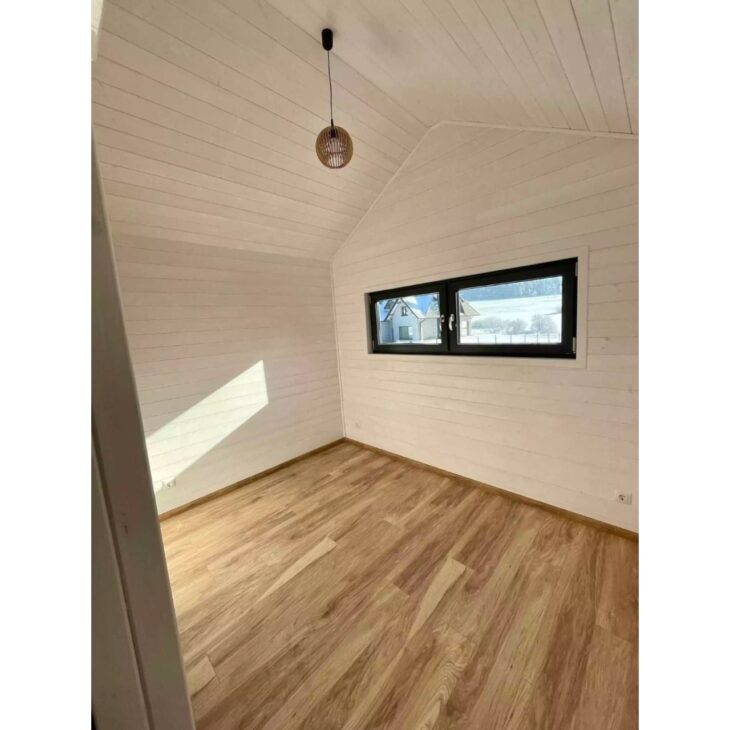 One of the bedrooms in the modular house shows how the project tries to escape the idea of cramped living, with wooden flooring, light walls, horizontal window, and free space to adapt the environment as a bedroom, office, or guest room.