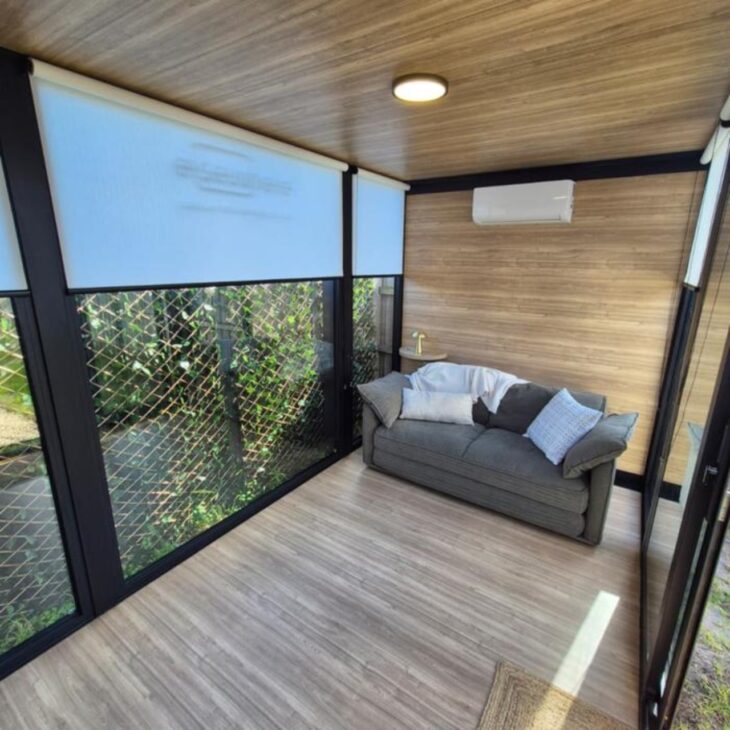 Interior do módulo desmontável vendido na Austrália mostra visual de cômodo pronto, com sofá, ar-condicionado e grandes janelas de vidro. O modelo menor custa cerca de R$ 94 mil, mede 2,7 m x 2,4 m e pode ser montado em poucos dias no quintal.