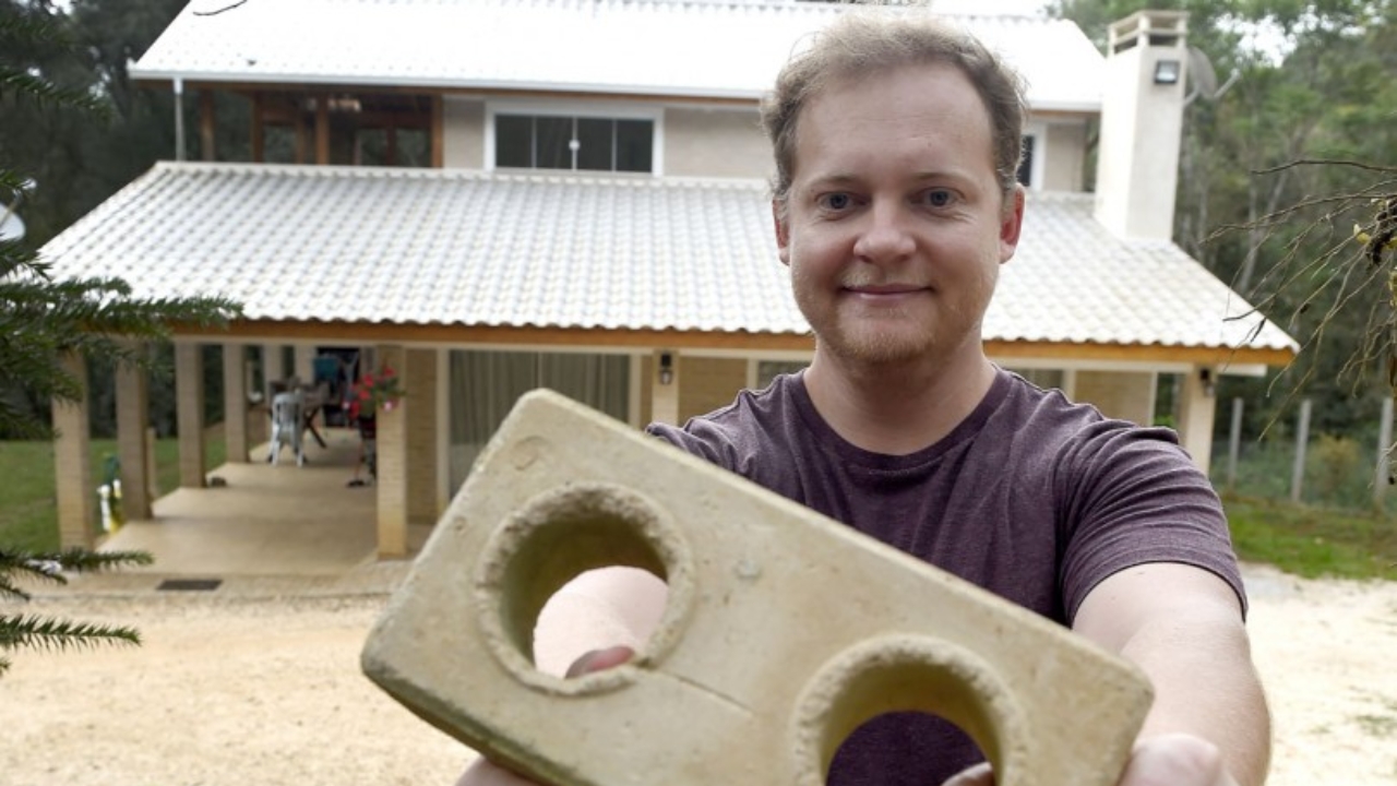 Com tijolo ecológico nas mãos e o sobrado ao fundo, o analista de sistemas mostra a casa de 200 m² erguida em Almirante Tamandaré após aprender etapas da obra com tutoriais no YouTube, reduzir custos e transformar um orçamento de R$ 12 mil apenas para a base estrutural em ponto de virada para construir praticamente por conta própria.