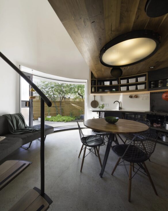 En el piso principal de la Silo House, cocina, sala y área de comedor comparten el mismo ambiente dentro de la antigua estructura circular. La apertura curva hacia el jardín, la escalera helicoidal, el acero negro y el techo revestido en madera muestran cómo el proyecto aprovechó los 31,6 m² sin crear divisiones que redujeran aún más el espacio.