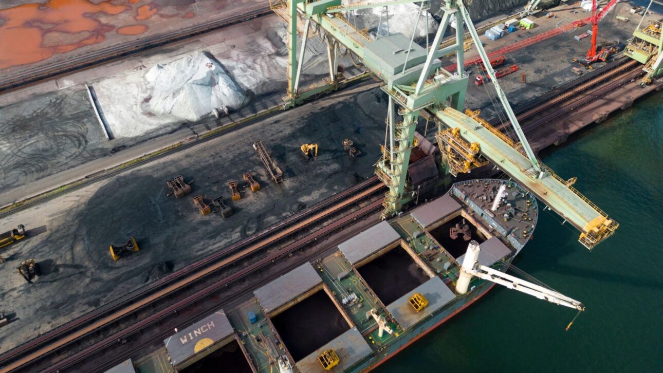 Navio graneleiro carrega minério de ferro em porto industrial, destacando a logística global ligada à demanda chinesa por aço.