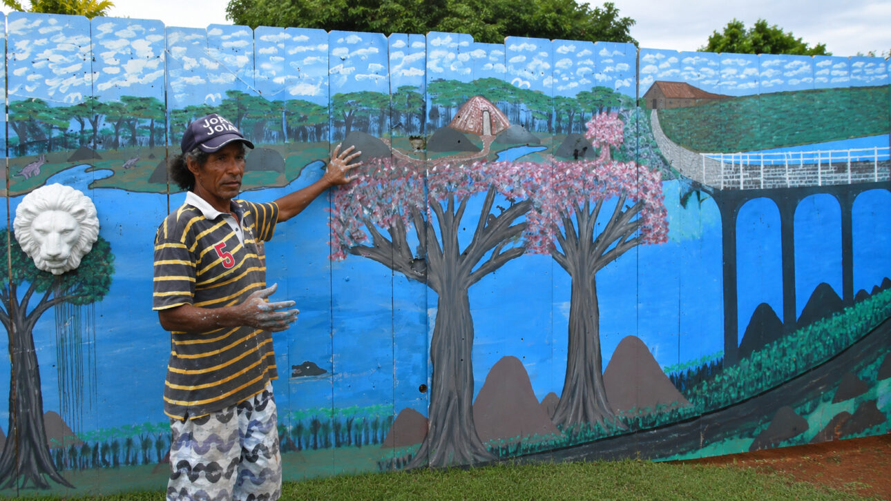 Homem em frente a muro azul com pintura artesanal de paisagem, árvores, ponte, rio e detalhes decorativos no jardim da casa.