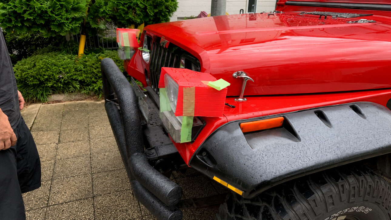 Red Jeep Wrangler with headlights temporarily repositioned on improvised cardboard and adhesive tape supports to meet Japanese vehicle inspection requirements.