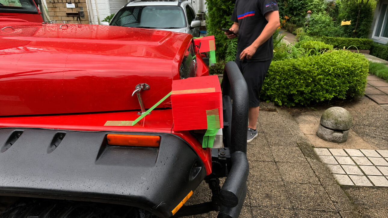 Front of red Jeep Wrangler with headlights mounted on cardboard boxes fixed with adhesive tape, a temporary adaptation used to meet Japanese vehicle inspection requirements.