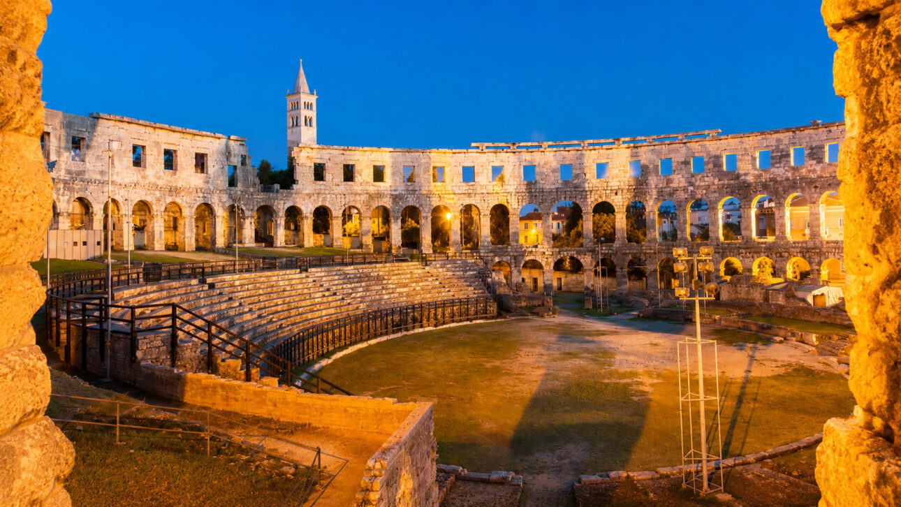 Interior do Anfiteatro de Pula ao entardecer, mostrando arquibancadas de pedra, arcos romanos preservados e a arena histórica localizada na Croácia.