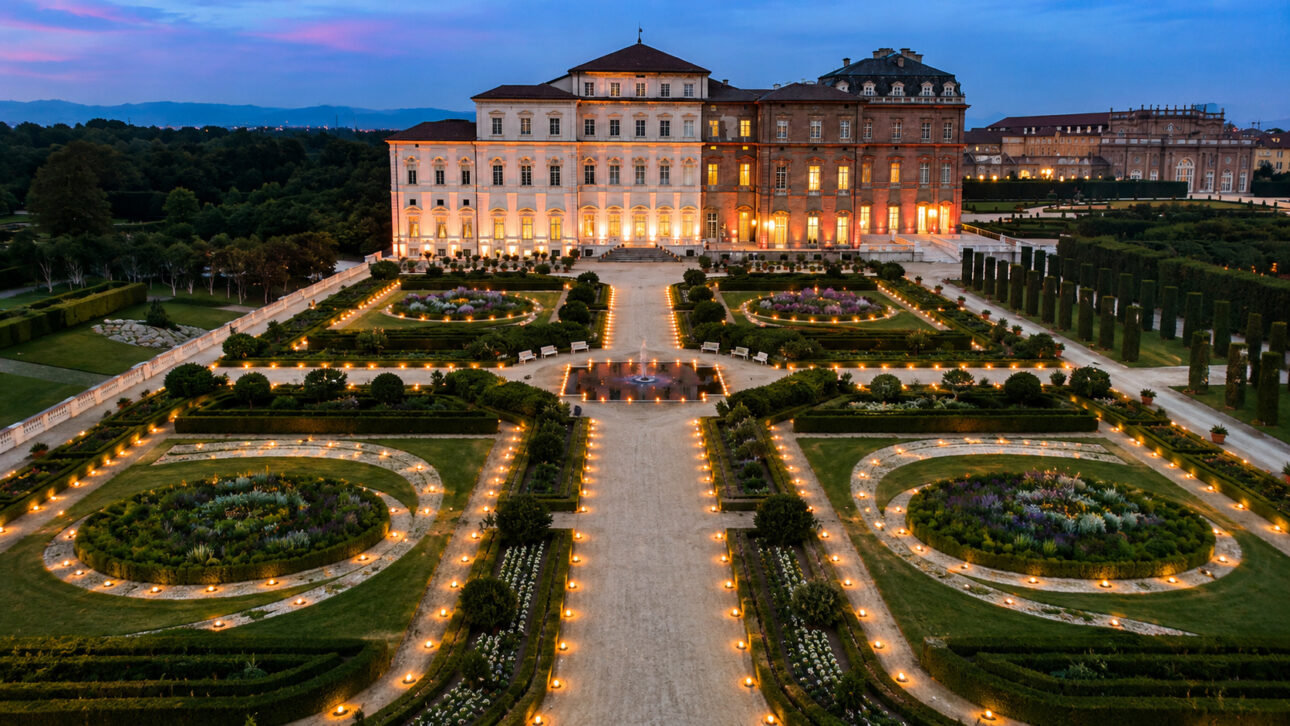 Panoramic view of the Reggia di Venaria Reale at sunset, highlighting the illuminated geometric Baroque gardens and the monumental royal residence of the House of Savoy in Piedmont, Italy.