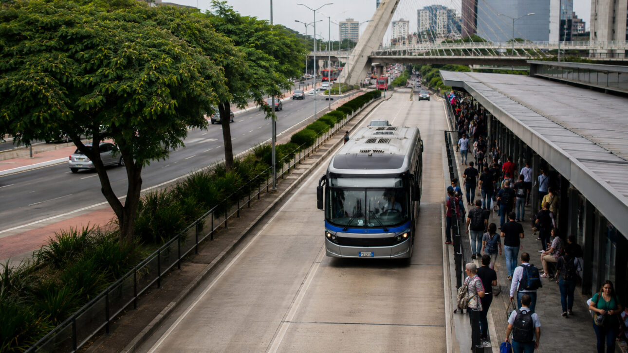 Ônibus moderno circula em corredor exclusivo ao lado de estação movimentada, ilustrando a mobilidade urbana e os impactos da nova lei do transporte público.