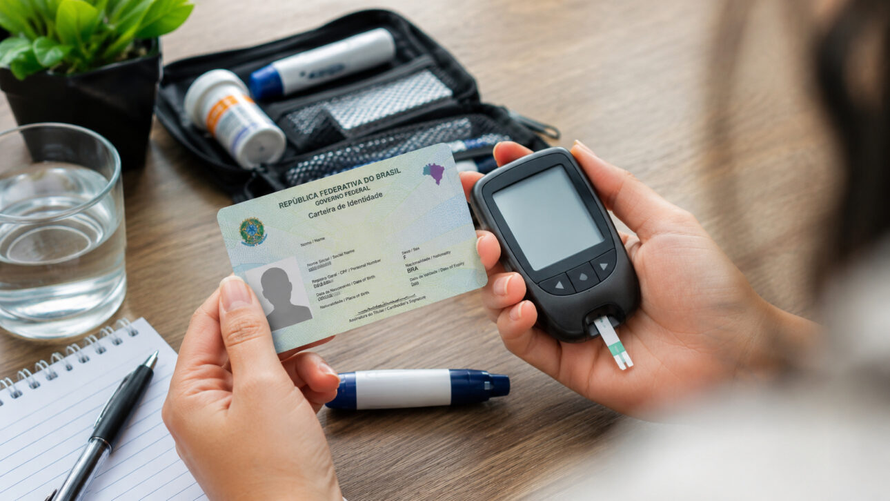 Mãos seguram um medidor de glicose e uma Carteira de Identidade Nacional sobre uma mesa com itens de monitoramento do diabetes tipo 1, ilustrando projeto que prevê identificação da condição no documento e novos direitos para pacientes.