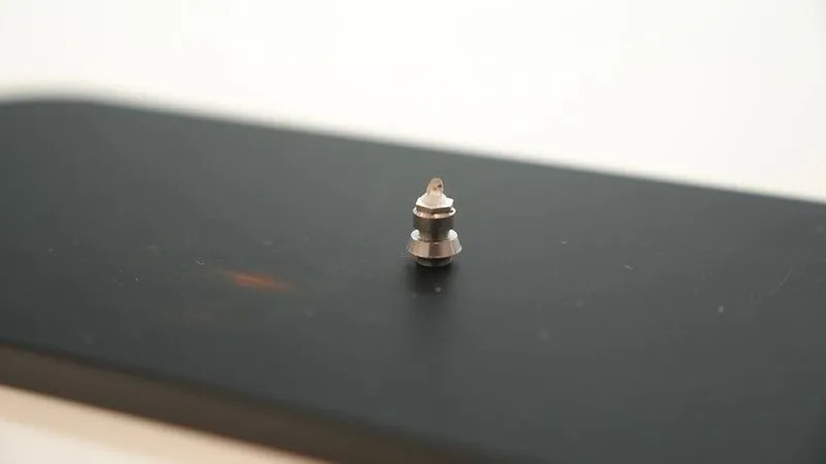 Small metallic piece in a conical shape positioned on a black surface, captured in close-up with a blurred light background.