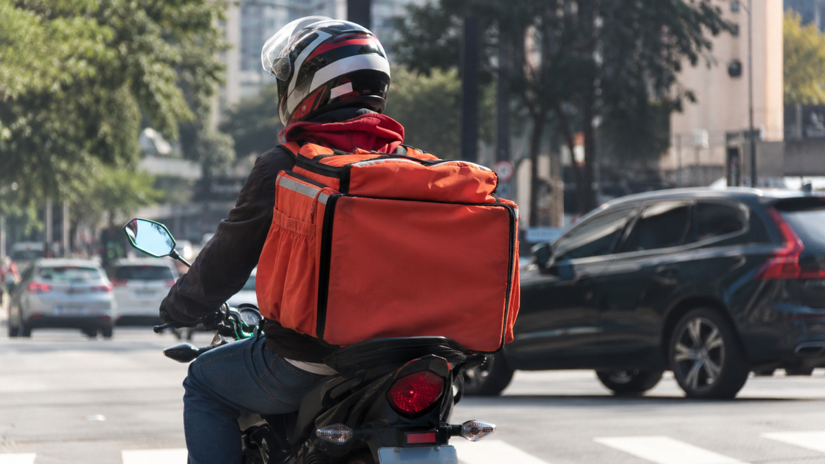 Entregador de aplicativo conduz motocicleta em área urbana movimentada, representando a proposta de crédito do governo para compra e substituição de motos usadas no trabalho.