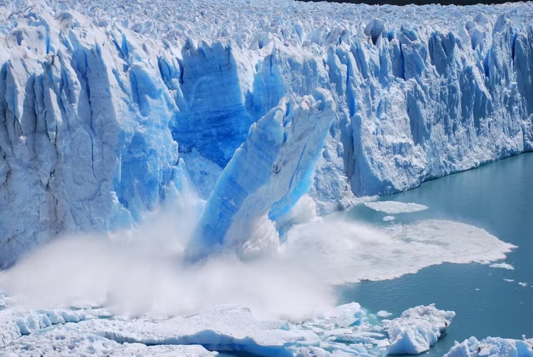 Bloco de gelo se desprendendo de uma geleira e caindo no oceano, formando ondas e fragmentos de gelo em ambiente polar associado ao aumento de icebergs na Groenlândia.