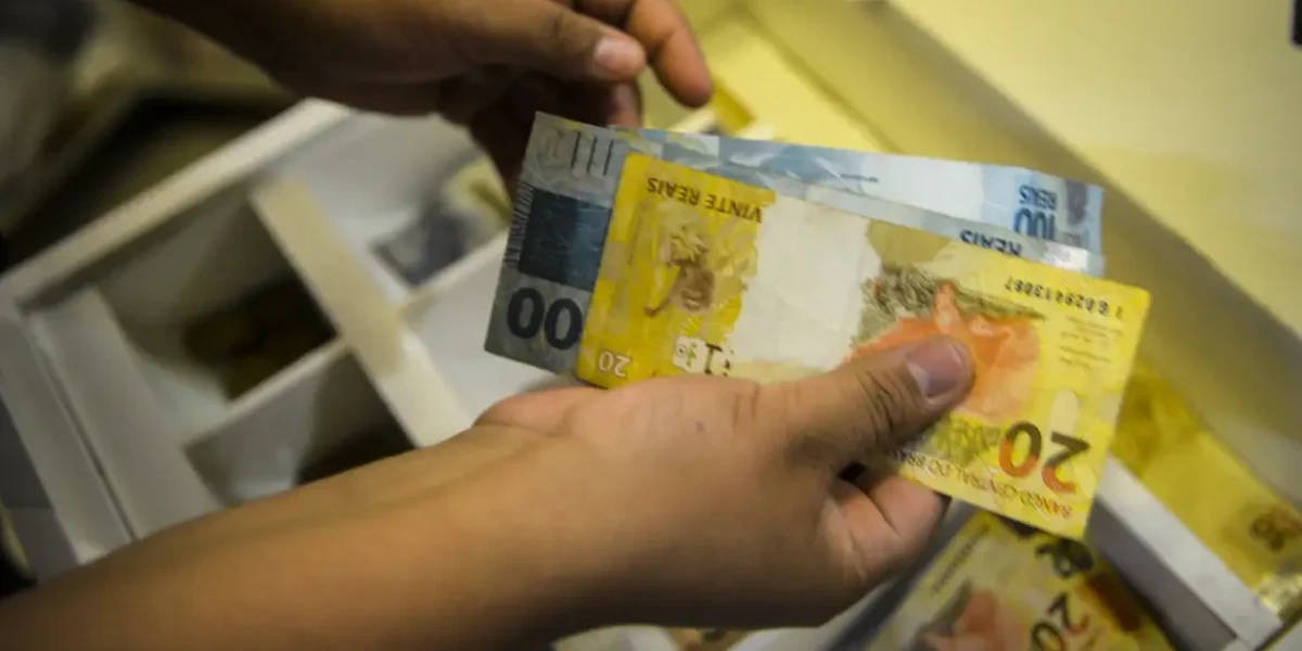 Person holding Brazilian real banknotes in front of a drawer with money, illustrating the payment of social benefits and the discussion about new rules for maintaining the BPC after entering the job market.