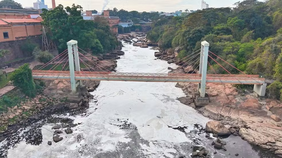 Imagen aérea muestra tramo del Río Tietê cubierto por espuma blanca entre áreas rocosas y un puente para peatones en Salto, en el interior de São Paulo, durante episodio asociado a la contaminación y a la reducción del caudal del río.