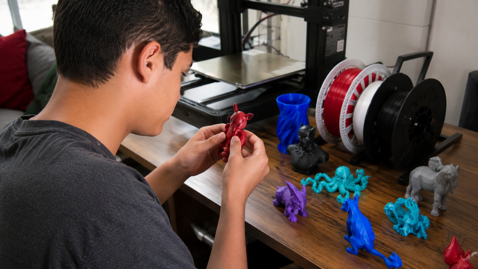 Adolescente analisa peça feita em impressora 3D sobre uma mesa com filamentos coloridos, miniaturas personalizadas e equipamento de impressão ao fundo.