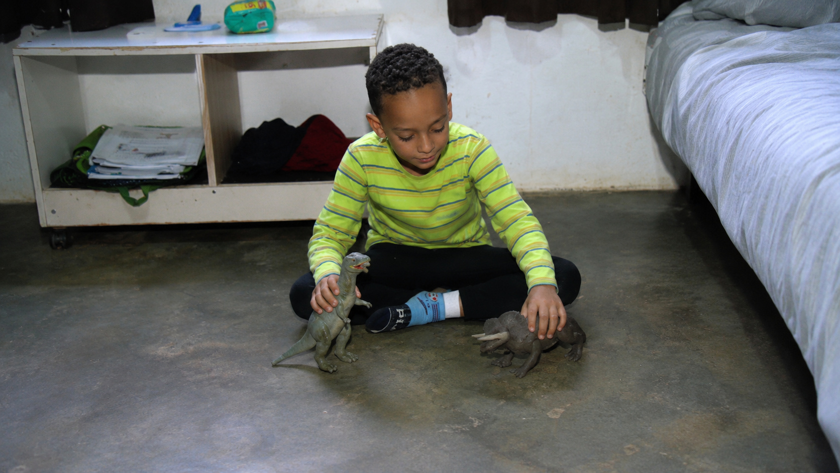 Menino sentado no chão do quarto brincando com dinossauros de brinquedo, em imagem ilustrativa sobre alergia rara à água e rotina adaptada.