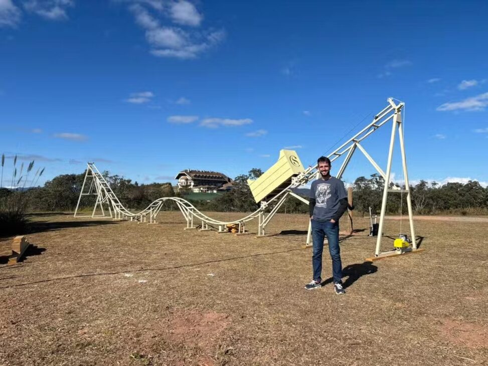 Jovem posa diante de uma montanha-russa construída em área aberta, com trilhos brancos, estrutura metálica e céu azul ao fundo.