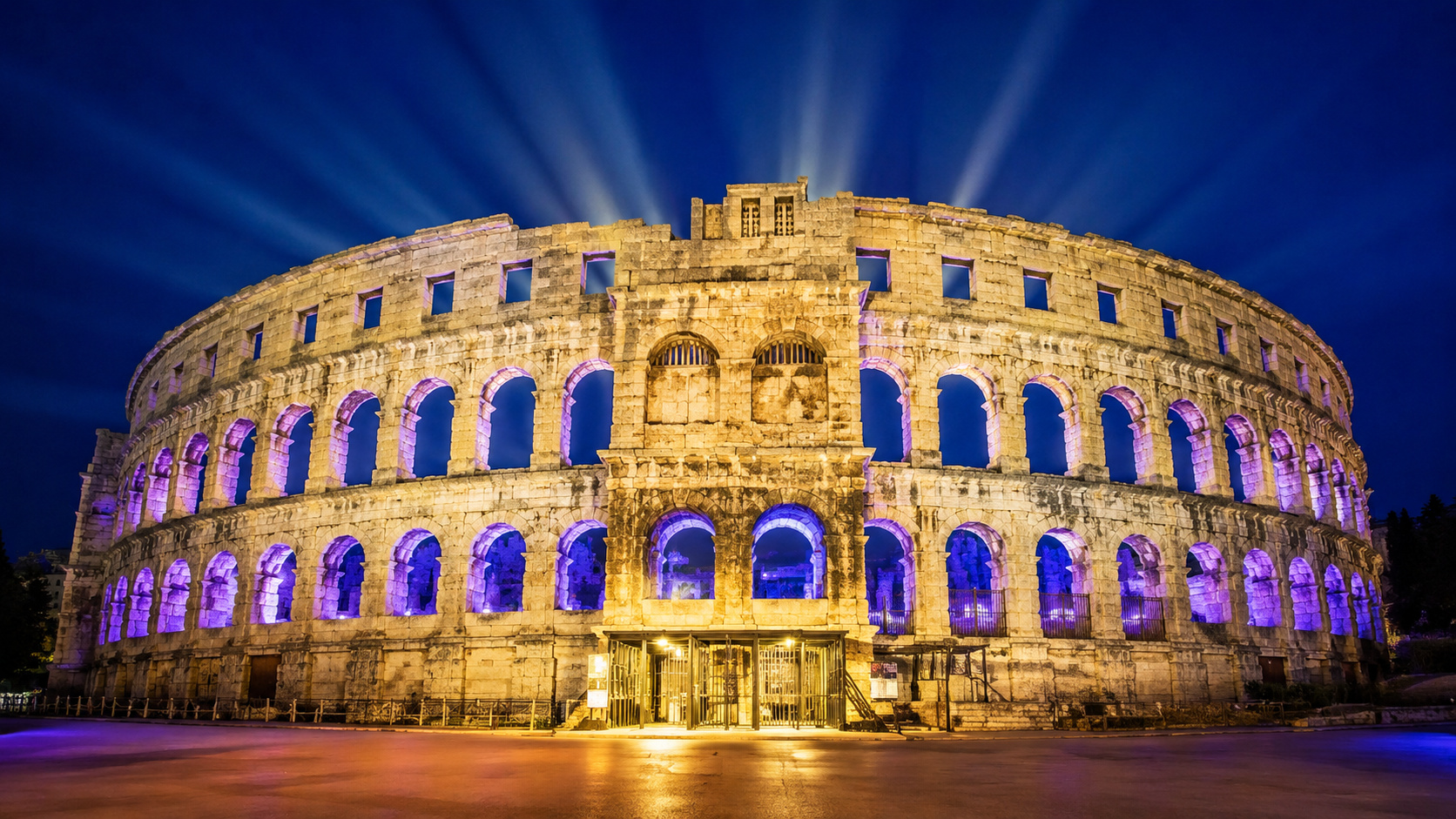 Anfiteatro romano iluminado durante a noite na Croácia, com muralhas preservadas, arcos monumentais e arquitetura do século I vista de frente.
