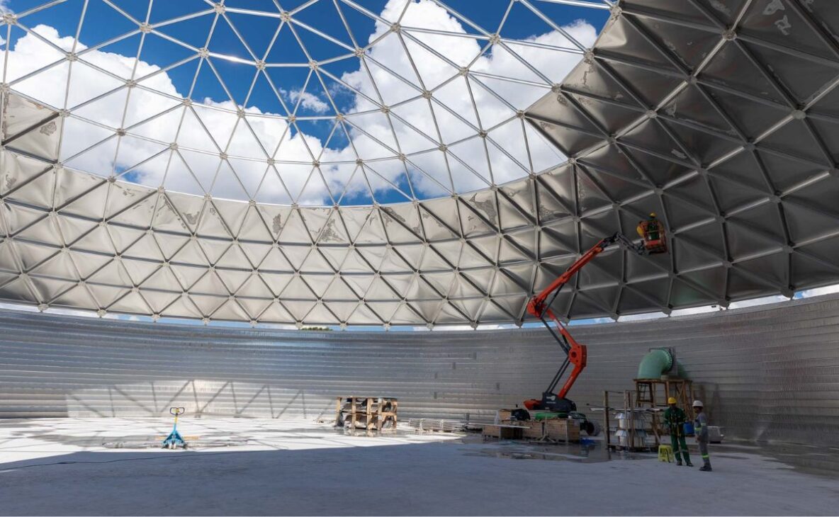 Vista interna do reservatório de água tratada construído com tecnologia de duplo aço, destacando a cobertura geodésica metálica, as paredes de aço inoxidável e os trabalhos de montagem realizados durante a construção da estrutura em Curitiba.