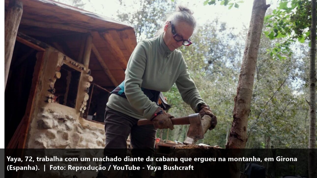 Yaya trabalhando com machado diante da cabana de madeira e pedra na floresta