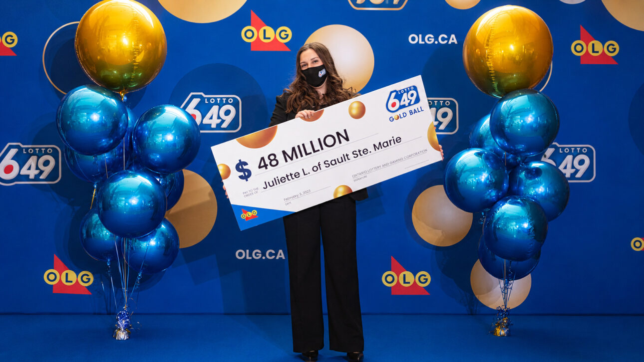 Joven sostiene un cheque de 48 millones de dólares canadienses en ceremonia de lotería, rodeada por globos azules y dorados.