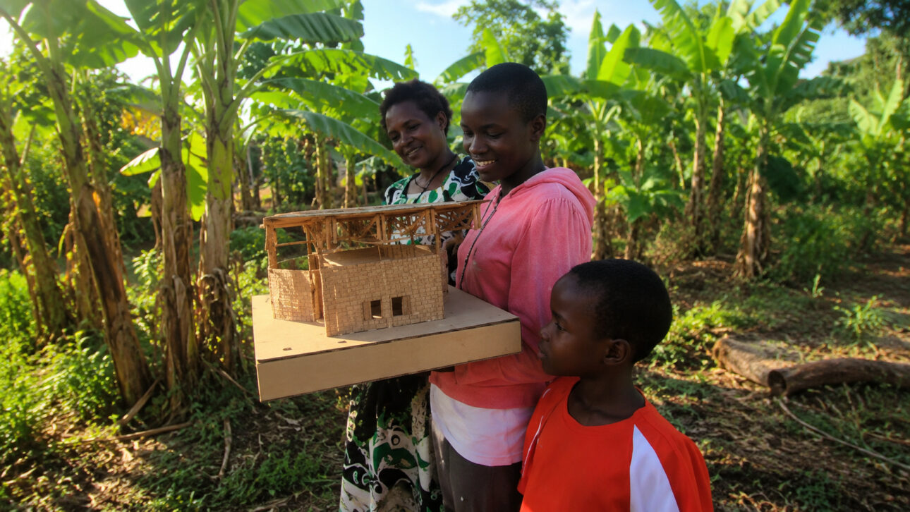 Mulheres e criança em área rural de Uganda observam maquete da Casa de Jajja cercados por bananeiras.