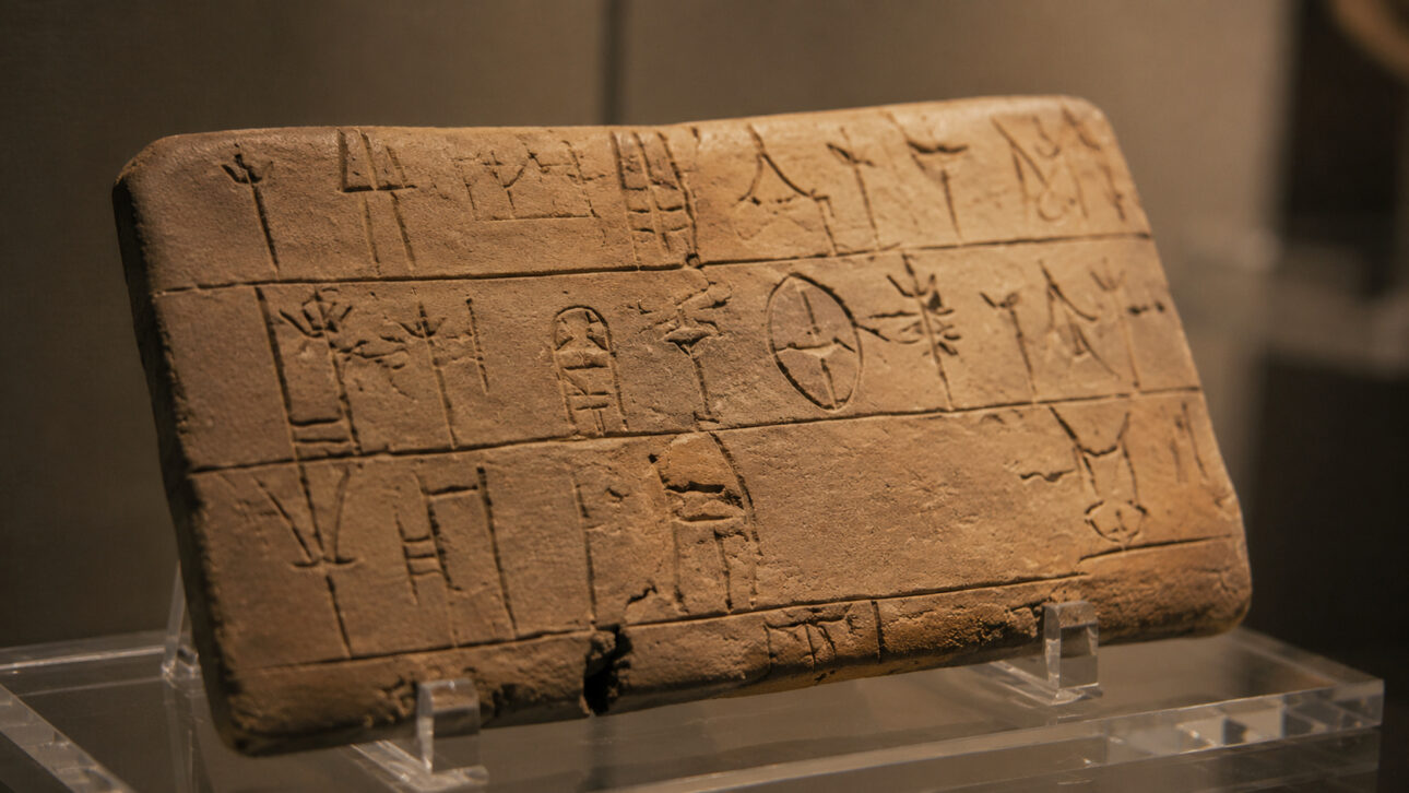Ancient clay tablet with undeciphered inscriptions displayed on a transparent stand inside a museum environment.