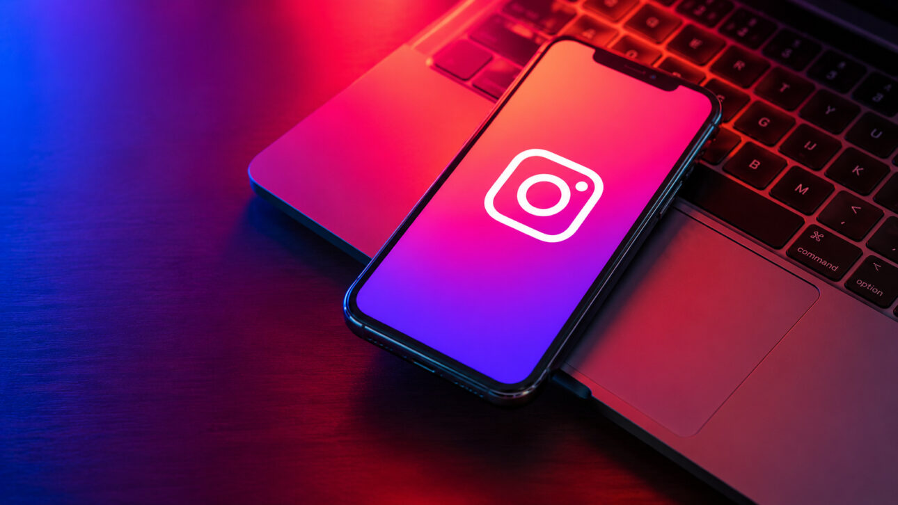 Cell phone displaying the Instagram icon on a colorful screen, with neon lighting and a notebook in the background.