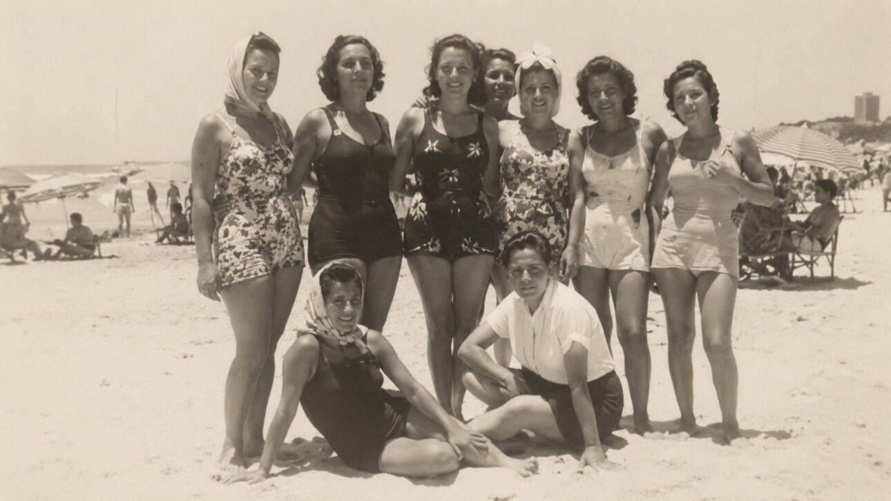 Fotografia em tom sépia mostra um grupo de mulheres com maiôs retrô reunidas na areia de uma praia, com guarda-sóis e banhistas ao fundo, representando a evolução da moda de praia antes da consolidação do biquíni.