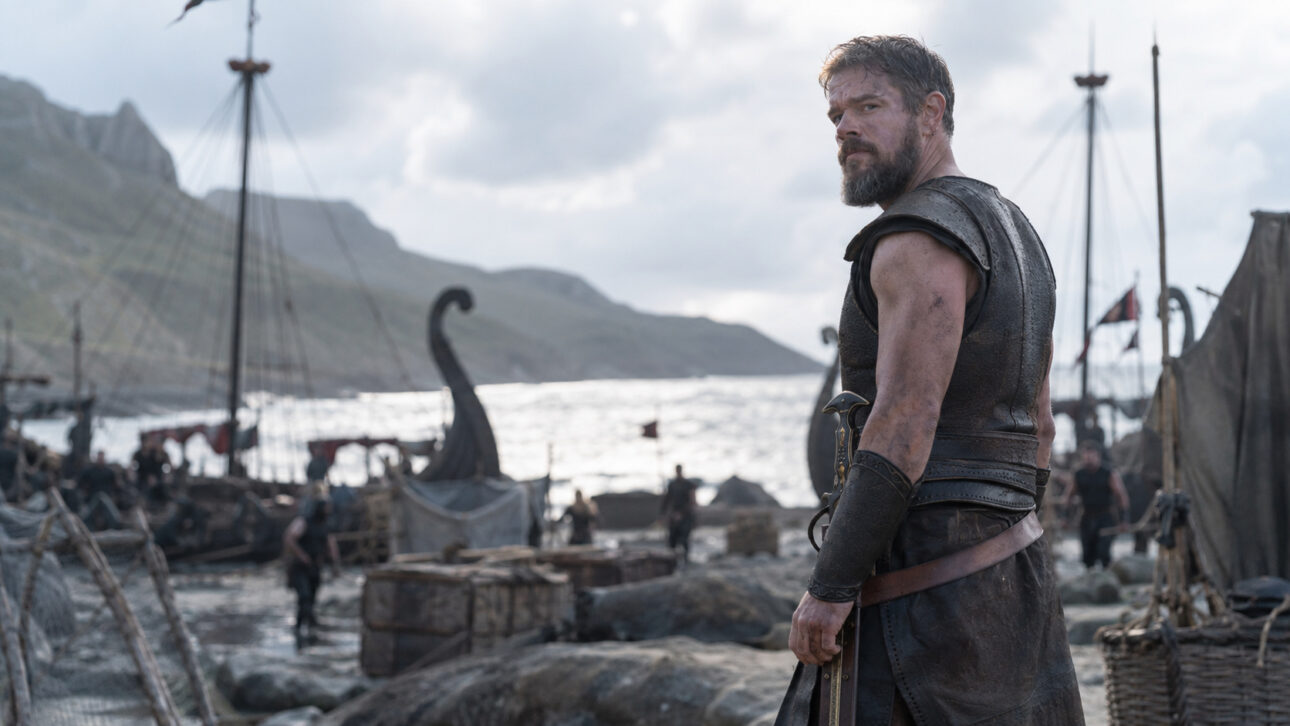 Viking warrior in the foreground in front of ships, tents, and a seaside camp under a cloudy sky.