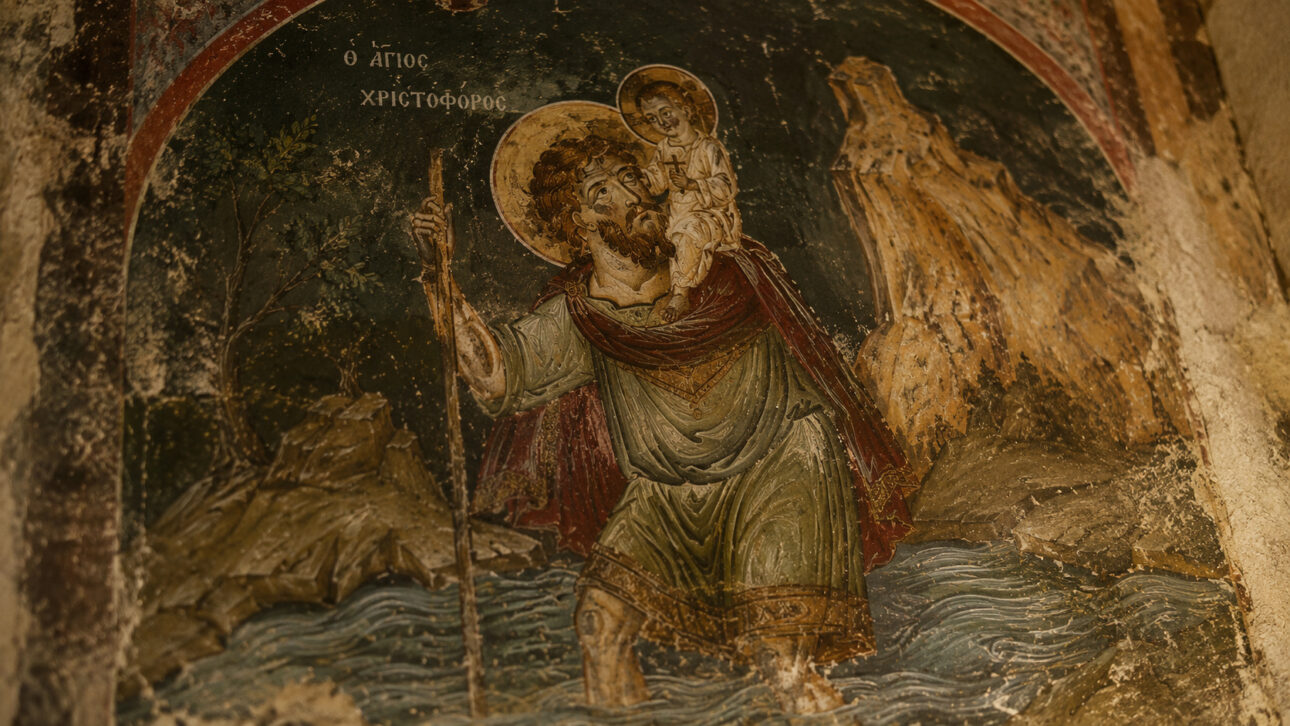 Afresco antigo de São Cristóvão carregando o menino Jesus nos ombros enquanto atravessa um rio com apoio de um cajado.