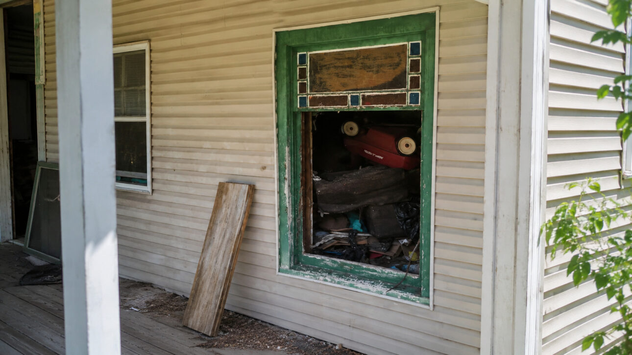 Janela de casa antiga com moldura verde descascada, objetos acumulados no interior e sinais de abandono relacionados ao caso das 16 crianças resgatadas em Ohio.