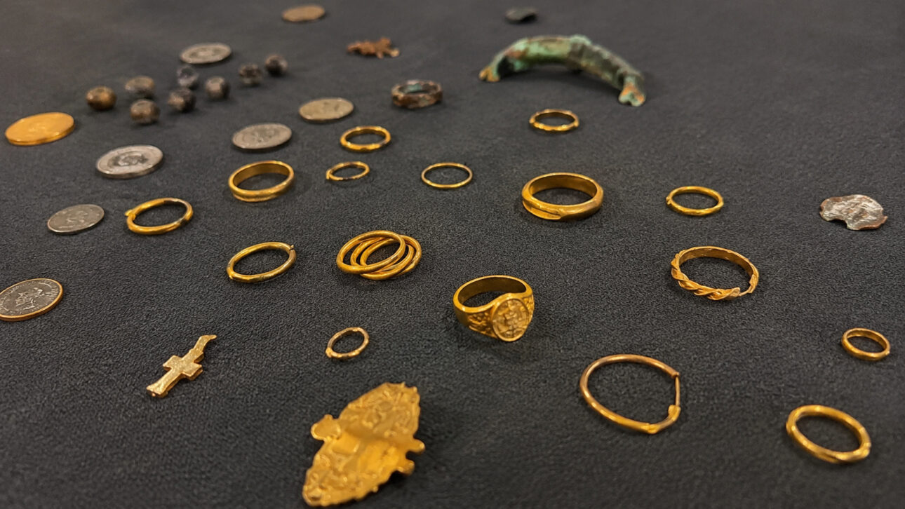 Joias de ouro, moedas antigas e fragmentos metálicos sobre tecido escuro, representando artefatos recuperados do navio Phoenix.