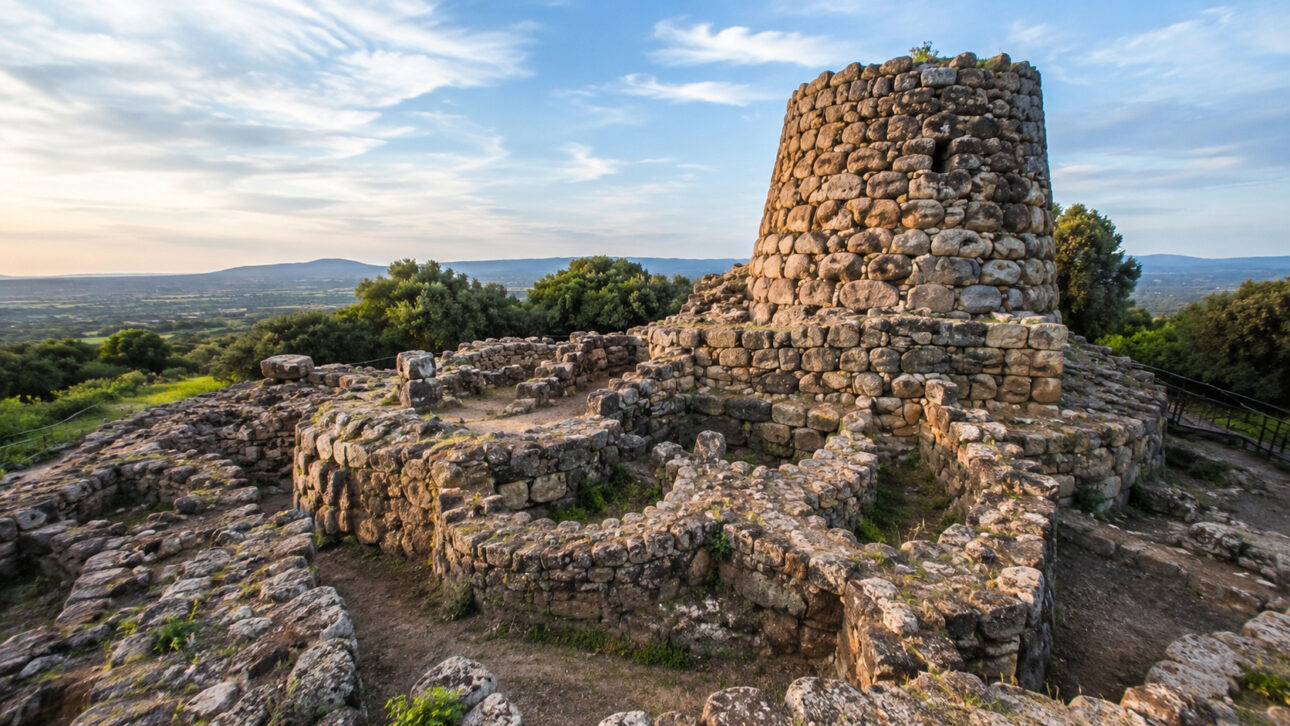 Ruínas de um nuraghe de pedra na Sardenha, com torre circular antiga, muralhas baixas e paisagem verde ao fundo.