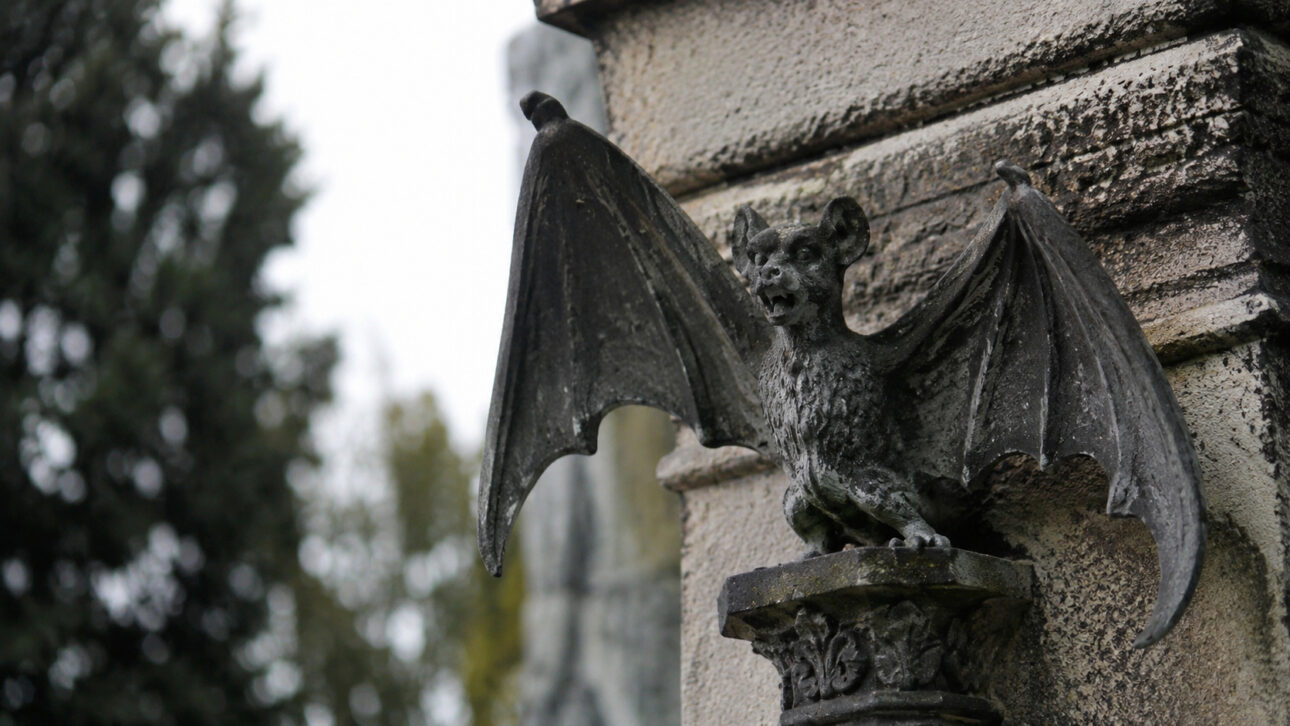 Escultura de morcego com asas abertas sobre estrutura de pedra em cemitério antigo, remetendo às lendas de vampiros do Père Lachaise.