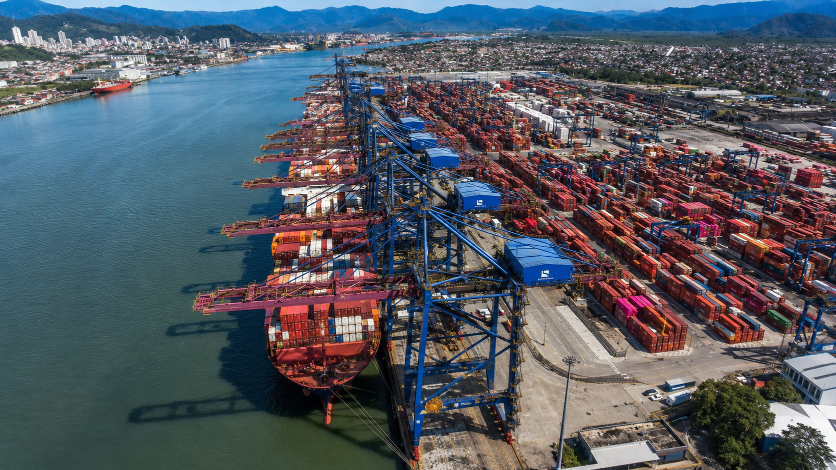 Vista aérea de terminal de contêineres no Porto de Santos, com navio transoceânico atracado, guindastes e cargas armazenadas.