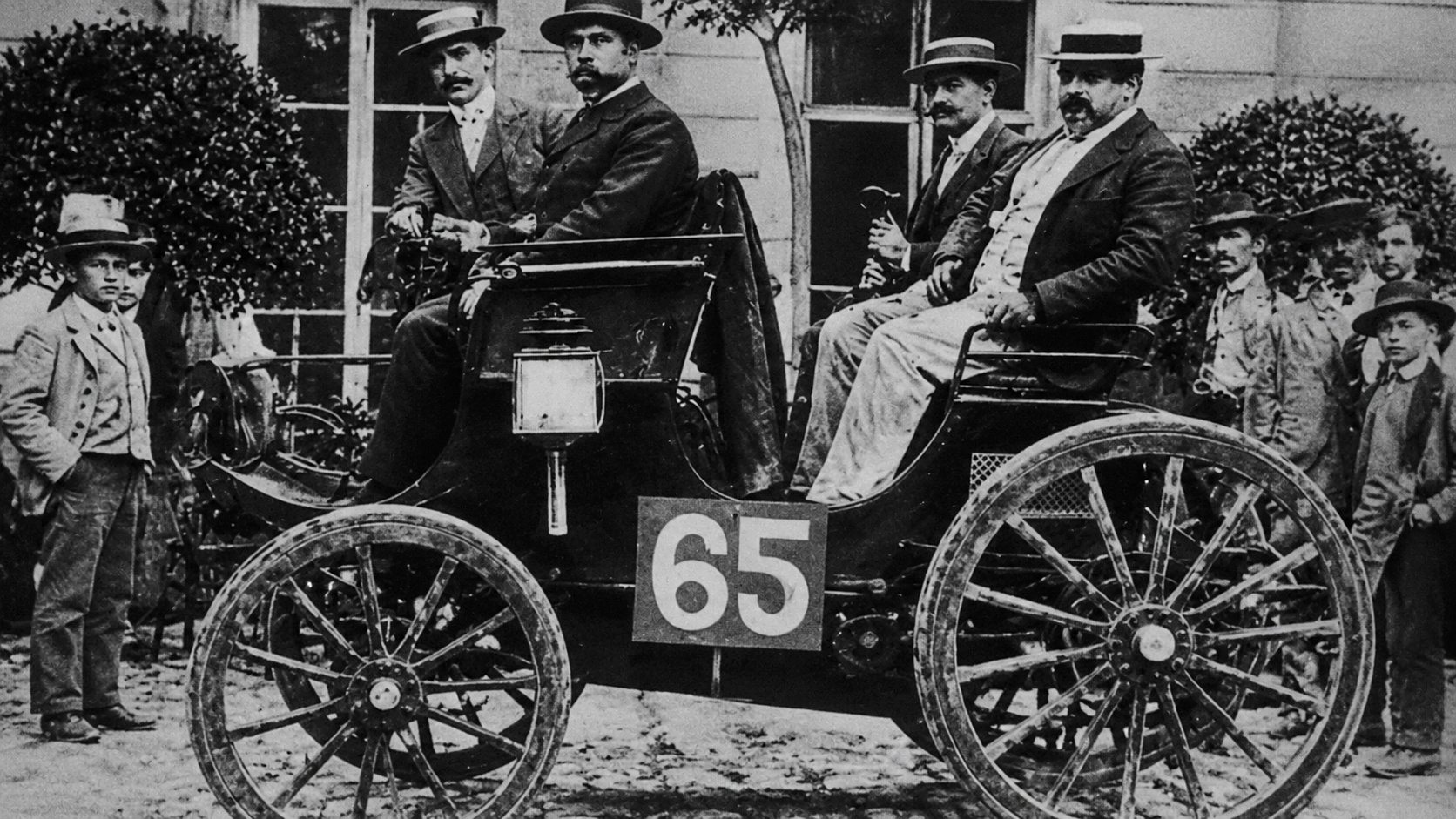 Automóvel da competição Paris-Rouen de 1894 com rodas de madeira e ocupantes em trajes da época, durante o evento que marcou a origem do automobilismo.