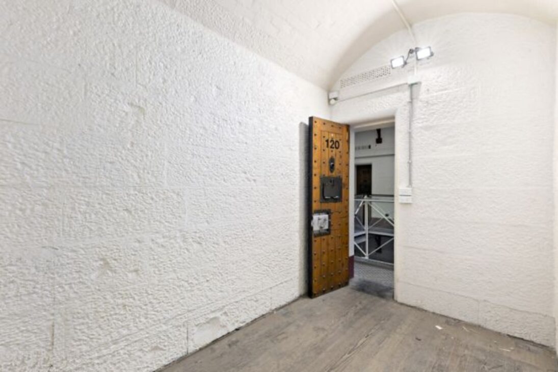 Interior da cela 120 com paredes espessas, porta de madeira reforçada e sistema de monitoramento de segurança.