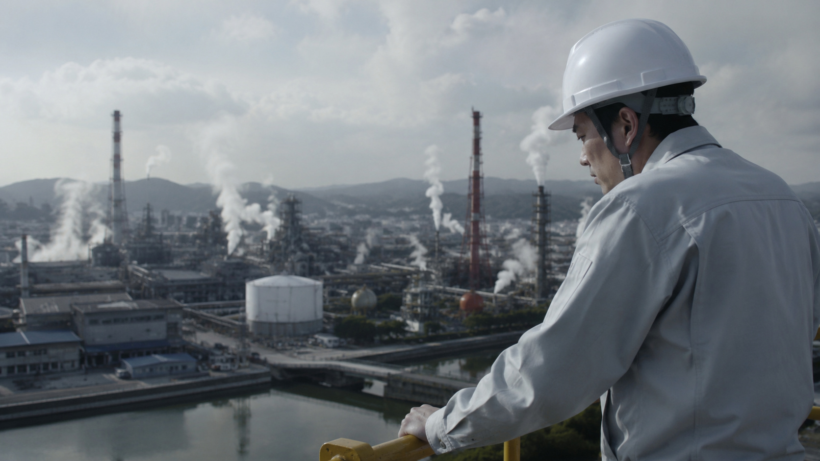 Trabalhador observa complexo industrial japonês com fábricas, chaminés e estruturas de produção ao fundo.