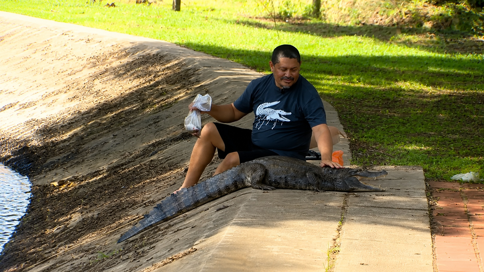 Homem sentado à beira de uma lagoa ao lado de um jacaré, em cena ilustrativa sobre a convivência entre Aldecino e Tião no Acre.