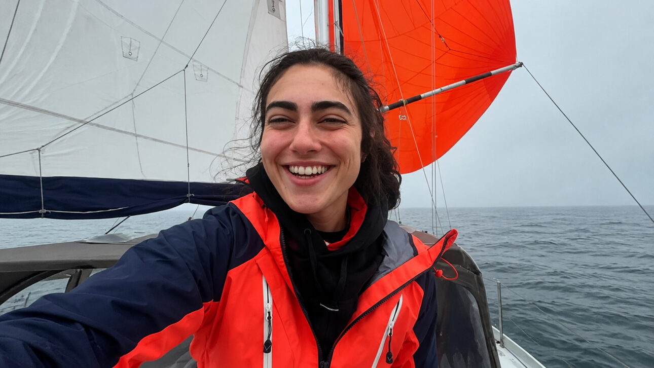 Mulher sorrindo com jaqueta de navegação em um veleiro, com velas abertas e o mar ao fundo.