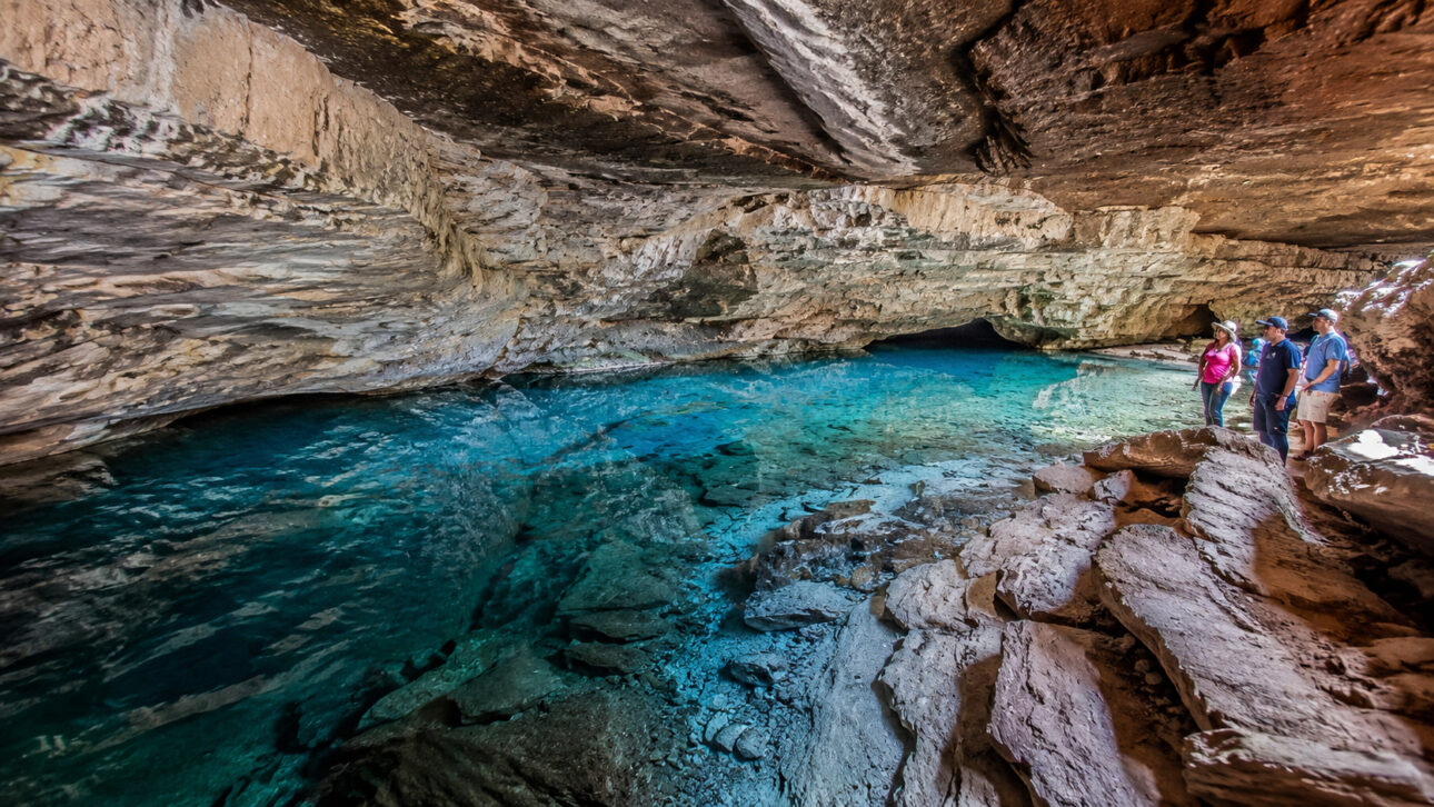 Interior de caverna com lago de água azul cristalina, paredes de calcário e visitantes em área turística de Iraquara, na Bahia.