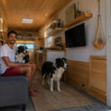 Homem dentro de caminhão adaptado como casa ao lado de dois cães, com móveis de madeira, cozinha, sofá e área de descanso.