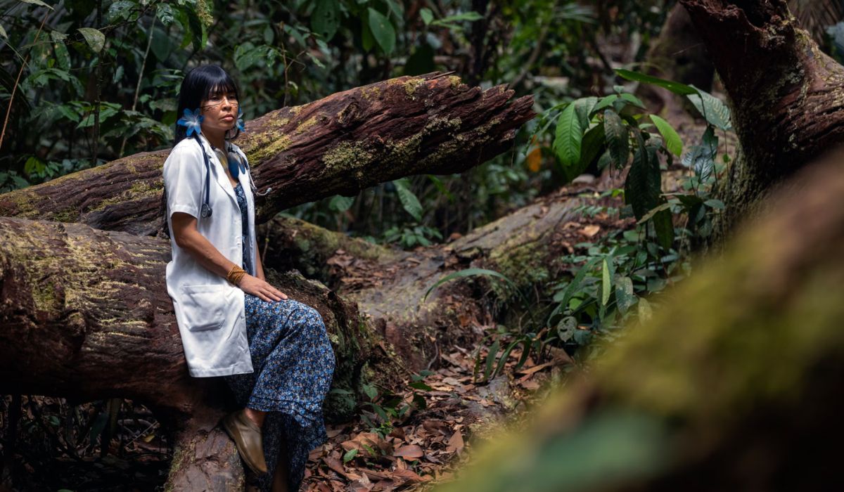 Com jaleco e estetoscópio em uma área de floresta, Adana representa visualmente o trabalho de aproximação entre o atendimento médico e as tradições de cuidado indígenas. Crédito: Divulgação/UFMG, via The Guardian.