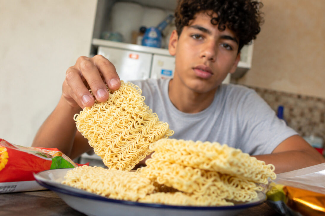 Adolescente segurando bloco de miojo cru diante de prato com macarrão instantâneo sem preparo, em imagem ilustrativa.